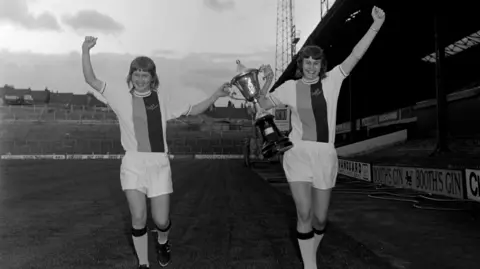 HY MONEY/ TOPFOTO Two Crystal Palace ladies players walking side-by-side on pitch holding a trophy together one hand each and their fists raised.