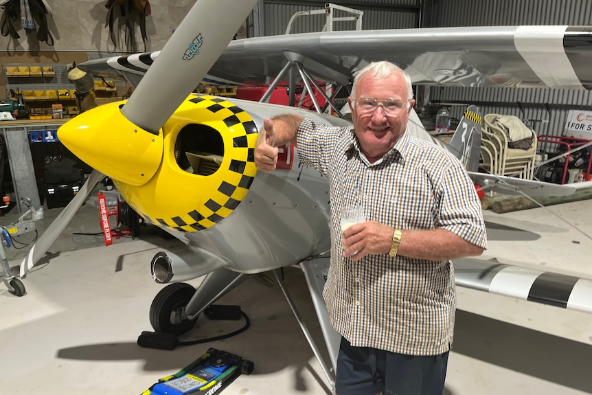 A man holding a glass of milk, standing beside a small aeroplane with yellow and grey colouring, inside a hanger. 