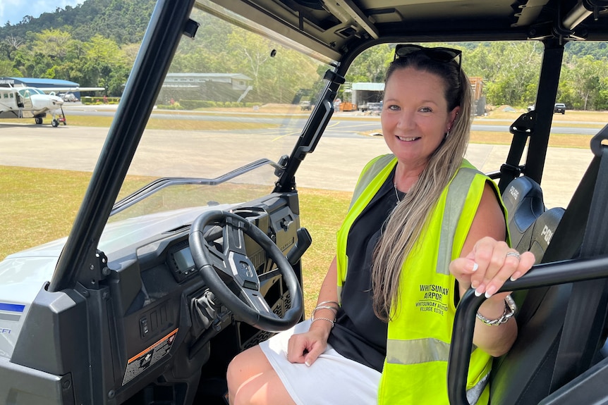  A woman in a hi-vis vest sitting in a buggy, on the edge of an airplane runway, with small planes in the background. 