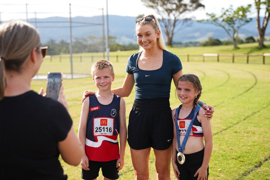 Jess with long blonde hair in a ponytail stands on a grass running track smiling with her arms around two young children. 